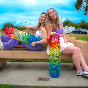 Two women holding rainbow stained glass skateboard decks at park.