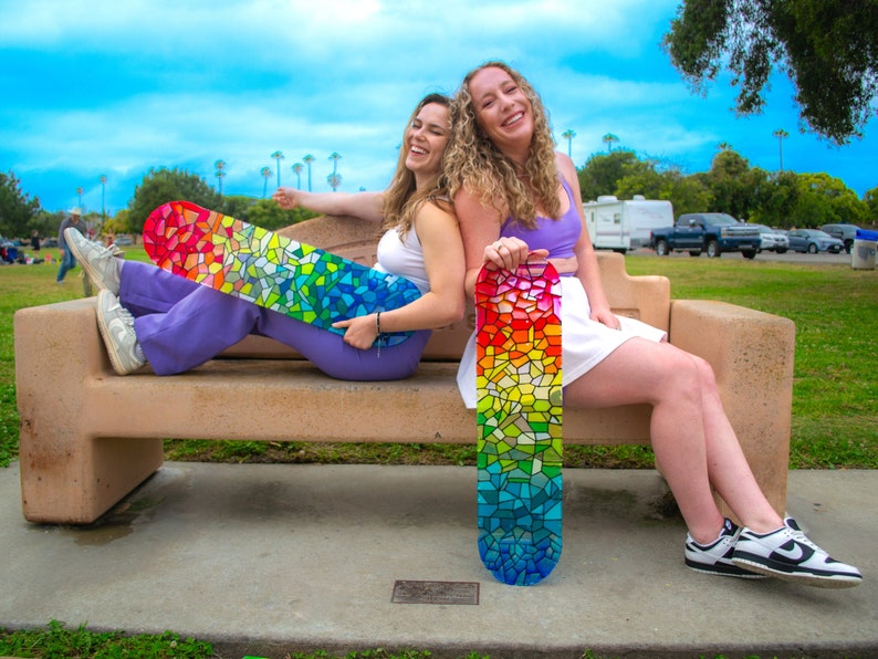 Two women holding rainbow stained glass skateboard decks at park.
