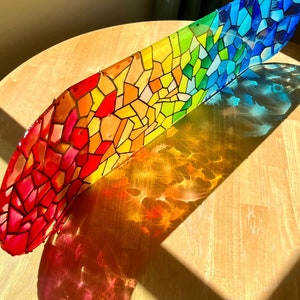 Rainbow stained glass skateboard deck artwork on table in sunlight casting colorful shadow.