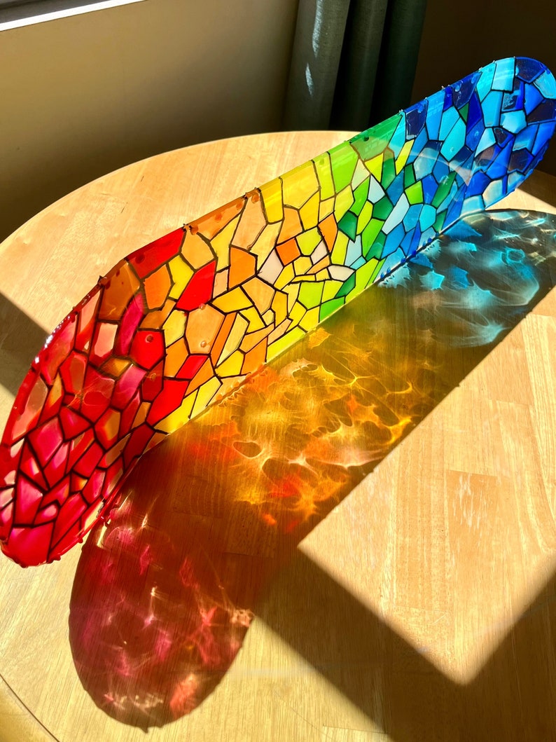 Rainbow stained glass skateboard deck artwork on table in sunlight casting colorful shadow.