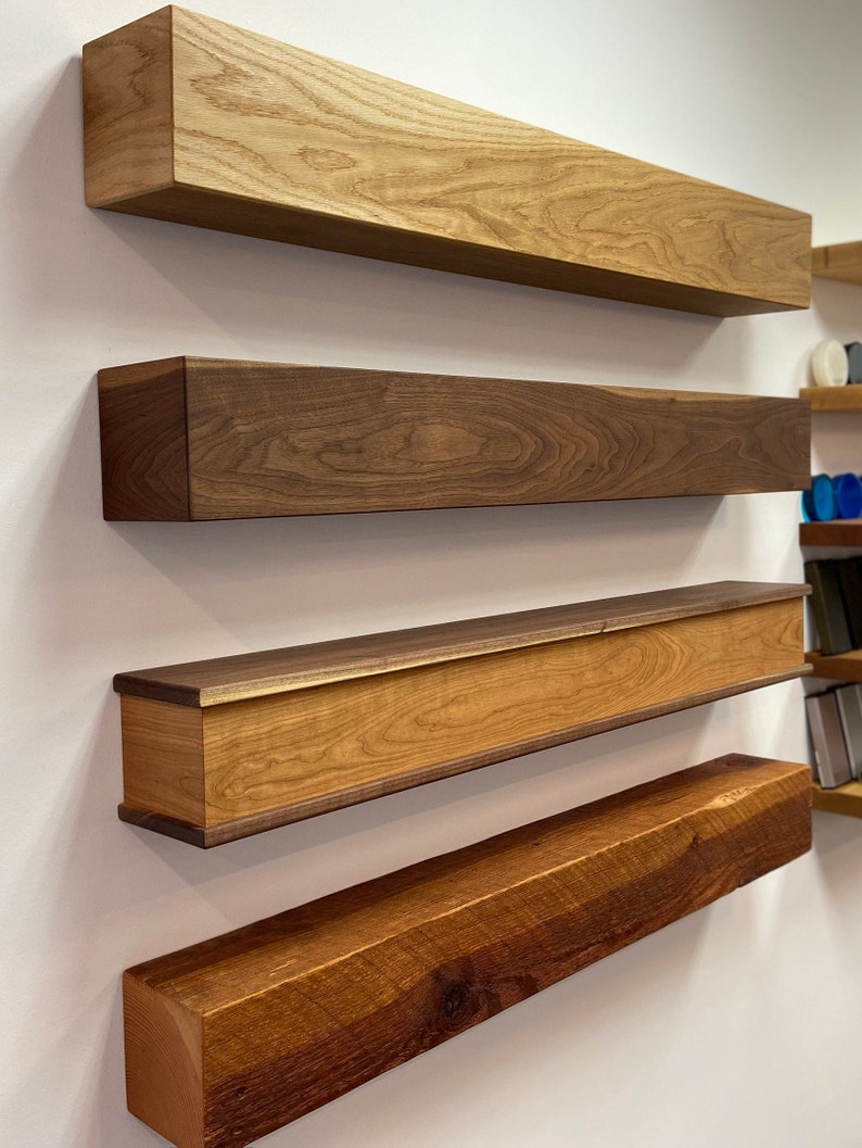 May include: Four wooden shelves, each made from a different type of wood, are mounted on a white wall. The shelves are all rectangular and have a smooth finish. The top shelf is a light brown colour, the second shelf is a dark brown colour, the third shelf is a reddish-brown colour, and the bottom shelf is a light brown colour with a rustic finish.