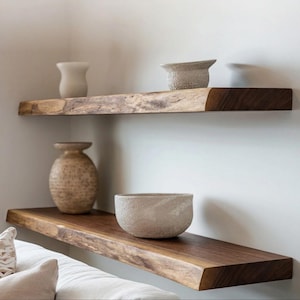 May include: Two horizontal floating shelves made of dark brown wood with natural live edges. The top shelf holds a small beige vase and a textured bowl. The bottom shelf holds a large, round, speckled tan vase and a wide, textured beige bowl.