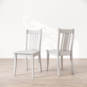 May include: Two white wooden chairs with a simple design. The chairs have a straight back with vertical slats and a slightly curved seat. The chairs are facing each other and are on a light wood floor.