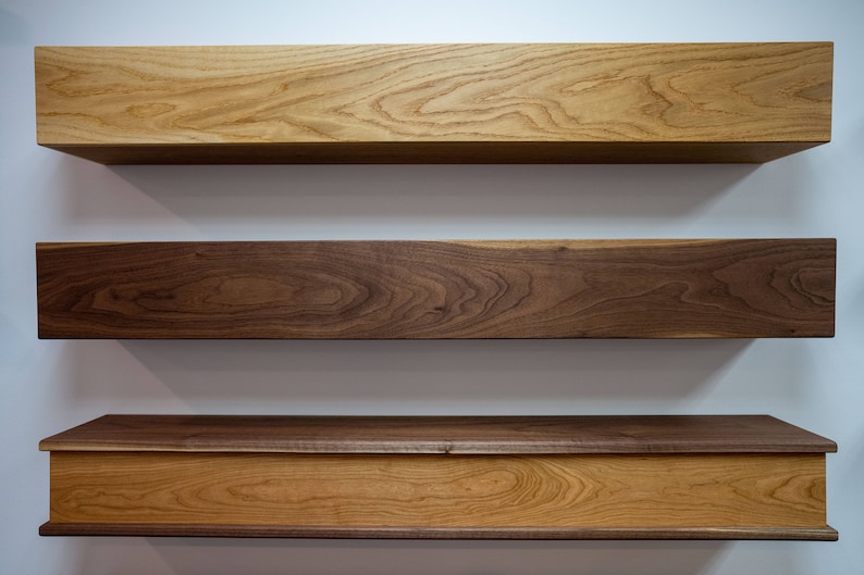 May include: Three wooden shelves, one with a drawer, mounted on a white wall. The top shelf is light wood, the middle shelf is dark wood, and the bottom shelf is light wood with a dark wood top.