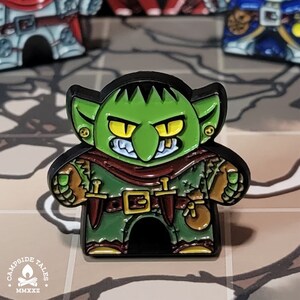 May include: A green goblin with a black and red outfit, holding a dagger and a pouch. The goblin has a toothy grin and yellow eyes. The pin is made of metal and has a black outline.