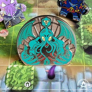 May include: A green and gold enamel pin featuring a Cthulhu-like creature with tentacles and a single eye. The pin is set against a background of a circular, ornate frame with a d20 die in the center.