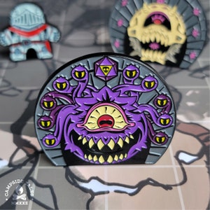 Peut inclure: Une épingle en émail violette et noire représentant un monstre béholder avec de multiples yeux et un grand œil central. L'épingle est sur un fond noir avec une bordure argentée et un dé d20 argenté.