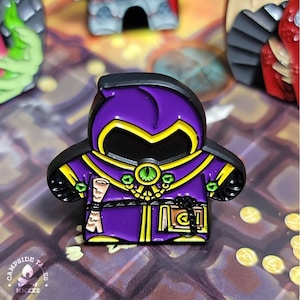 May include: Purple and gold enamel pin of a wizard with a scroll and a book. The wizard is wearing a purple robe with a gold hood and a gold belt. The pin is on a black and yellow background.