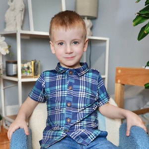 May include: A young boy wearing a blue and green plaid shirt with a white collar and blue jeans. He is sitting on a white chair with a blue cushion.