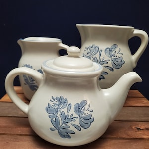 Puede incluir: Un juego de tres piezas de cocina de cerámica blanca con diseños florales azules. El juego incluye una tetera, una jarra y una jarrita.