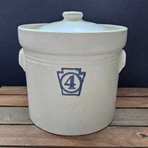 May include: White ceramic crock with a lid, featuring a blue Pennsylvania state outline with the number "4" inside. The crock is sitting on a wooden surface.