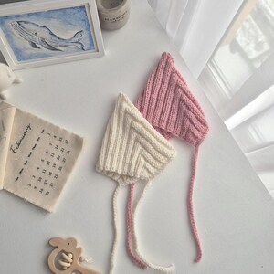 May include: Two knitted baby bonnets, one cream and one pink, with long ties, rest on a white surface. A wooden baby rattle and a framed whale painting are also visible. A small calendar shows the month of February.