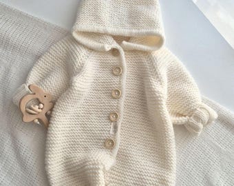 May include: Cream-colored knitted baby jumpsuit with a hood and wooden buttons. The jumpsuit has long sleeves and legs, and is paired with a wooden bunny-shaped teether. A white ceramic bunny figurine is in the background.