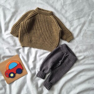 Pode incluir: Um suéter de malha marrom e uma calça de malha cinza para um bebê. O suéter tem uma textura canelada e a calça tem uma cintura com cordão.