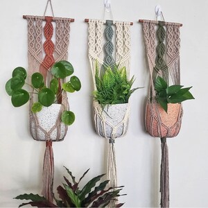 May include: Three macrame plant hangers in beige, brown, and green, each holding a speckled white or peach-coloured pot with greenery. The hangers are suspended from wooden dowels and feature intricate knotting patterns, adding a bohemian touch to the decor.