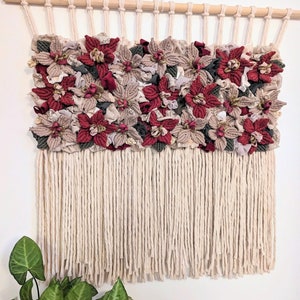 May include: A macrame wall hanging with a wooden dowel. The woven section features burgundy and beige fabric flowers with gold accents. Cream-coloured fringe hangs below the floral design, creating a textured, decorative piece for the home.