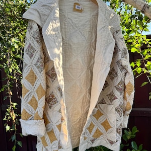May include: Cream-colored quilted jacket with a hood and open front. The jacket features a diamond pattern in shades of cream and gold, with floral accents on the sleeves. The jacket is hanging on a wooden hanger against a backdrop of greenery.