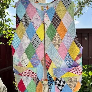 May include: A colorful patchwork vest with a diamond pattern, featuring various fabrics in shades of pink, blue, yellow, green, and orange. The vest has two pockets and is hanging on a wooden hanger against a natural background.
