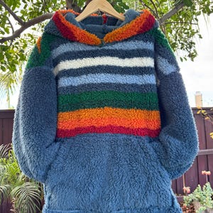 May include: A blue fleece hoodie with a colorful striped design across the chest. The stripes include red, orange, green, white, and blue. The hood trim is orange and red. The hoodie is hanging on a wooden hanger.