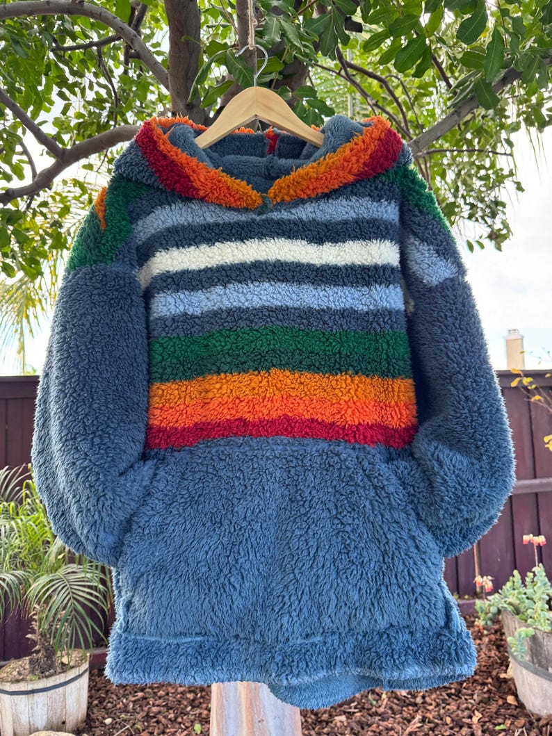 May include: A blue fleece hoodie with a colorful striped design across the chest. The stripes include red, orange, green, white, and blue. The hood trim is orange and red. The hoodie is hanging on a wooden hanger.