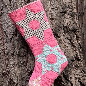 May include: A quilted Christmas stocking featuring a pink fabric base with star-shaped patchwork patterns. The upper star displays a black and white checkered design, while the lower star features a light blue floral print with pink accents.