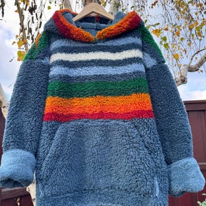 May include: A blue, hooded sweatshirt with a kangaroo pocket and striped design. The stripes include white, green, orange, and red. The hood and cuffs have a textured, colorful trim. The sweatshirt is hanging on a wooden hanger.