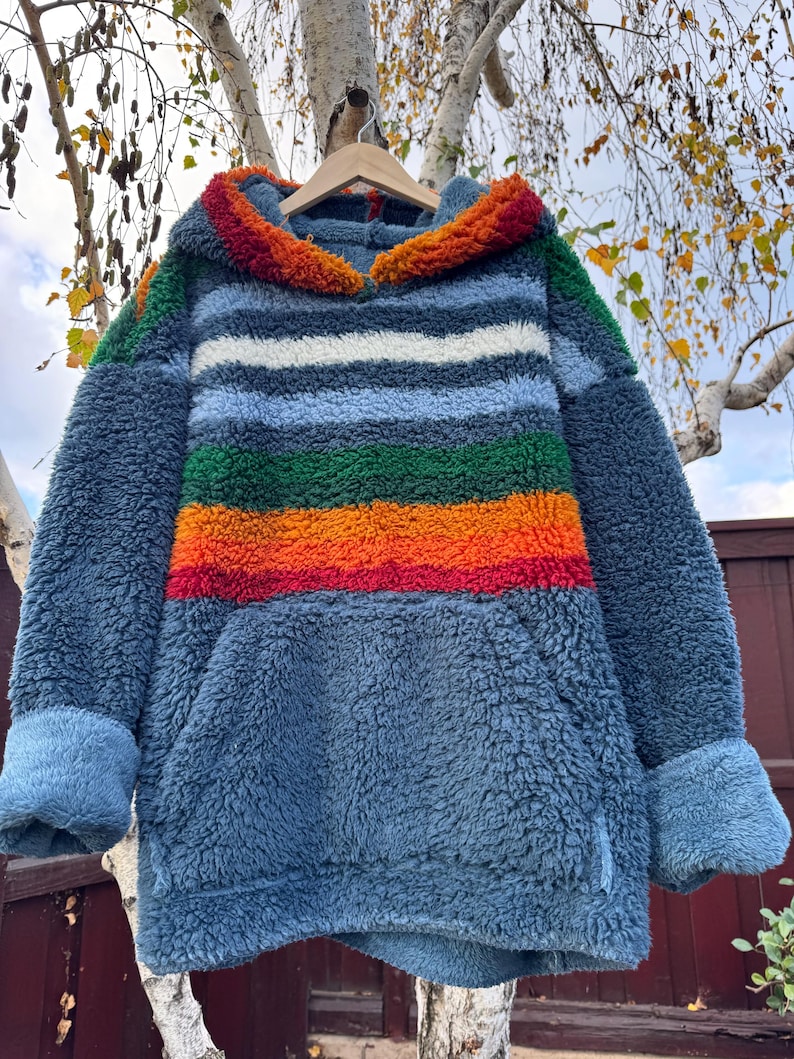 May include: A blue, hooded sweatshirt with a kangaroo pocket and striped design. The stripes include white, green, orange, and red. The hood and cuffs have a textured, colorful trim. The sweatshirt is hanging on a wooden hanger.