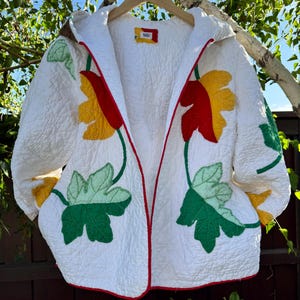 May include: White quilted jacket with a hood, adorned with colorful leaf appliqués in green, yellow, and red. The jacket features a red trim and is hanging on a wooden hanger. The jacket is a unique piece of outerwear.