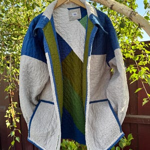 May include: A quilted jacket with a geometric color block design. The jacket features shades of blue, green, and gray, with a textured fabric. The jacket is hanging on a wooden hanger against a backdrop of green foliage and a wooden fence.