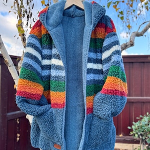 May include: A blue hooded sherpa robe with rainbow striped sleeves and a blue interior. The robe has two pockets and is hanging on a wooden hanger. The robe is made of a soft, plush material.