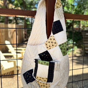 May include: Cream-colored quilted shoulder bag with patchwork squares in black floral, green, and yellow patterns. The bag has a large pocket and a wide strap. The bag is hanging on a wooden post.