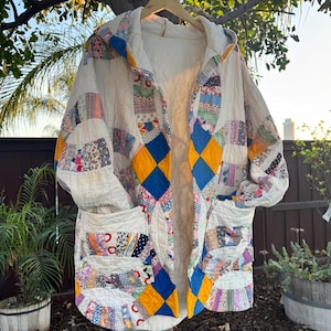 May include: A handmade, quilted coat with a hood, featuring a patchwork design in various colors and patterns. The coat has a cream-colored base with blue and yellow diamond accents, and two front pockets. It is hanging on a wooden hanger outdoors.