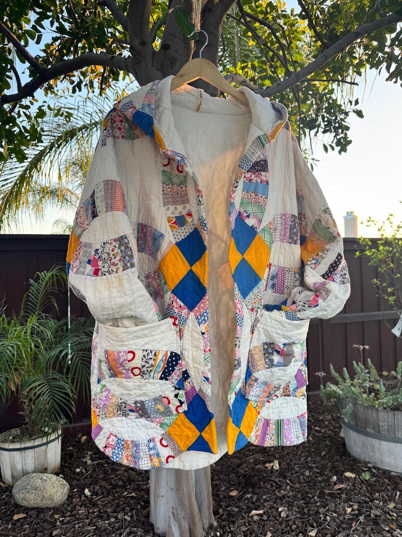 May include: A handmade, quilted coat with a hood, featuring a patchwork design in various colors and patterns. The coat has a cream-colored base with blue and yellow diamond accents, and two front pockets. It is hanging on a wooden hanger outdoors.