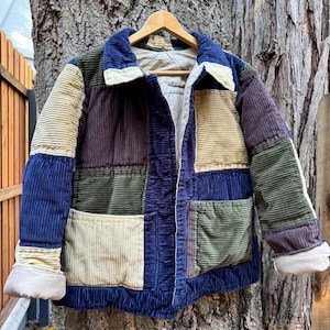 May include: A patchwork corduroy jacket in shades of blue, green, tan, and purple. The jacket has a collared neckline, two front pockets, and cream-colored cuffs. It is hanging on a wooden hanger against a tree trunk.