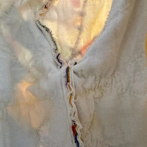 May include: Close-up of a white quilted fabric with a raw edge. The edge is adorned with colorful threads in shades of red, blue, yellow, and green. The fabric has a soft, textured appearance.