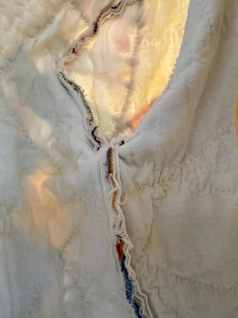 May include: Close-up of a white quilted fabric with a raw edge. The edge is adorned with colorful threads in shades of red, blue, yellow, and green. The fabric has a soft, textured appearance.