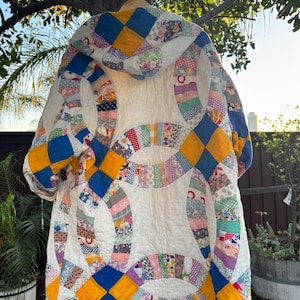 May include: A handmade, vintage quilt coat with a unique design. The coat features a white background with colorful patchwork circles and diamond patterns in blue, yellow, and other colors. The coat is hanging from a wooden hanger.