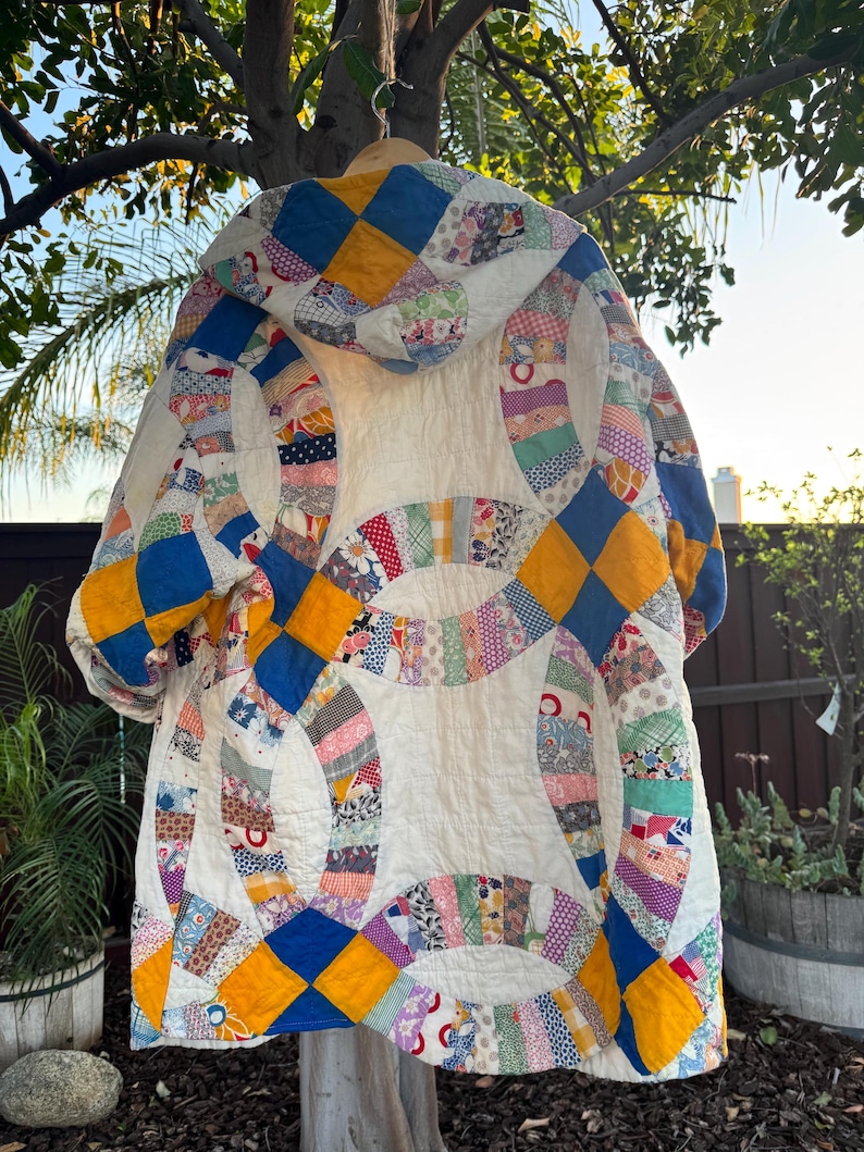 May include: A handmade, vintage quilt coat with a unique design. The coat features a white background with colorful patchwork circles and diamond patterns in blue, yellow, and other colors. The coat is hanging from a wooden hanger.