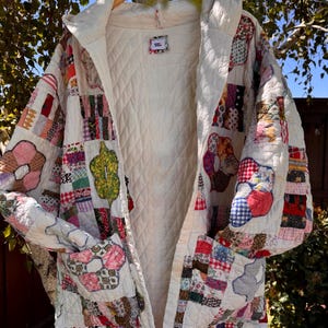 May include: A quilted jacket featuring a patchwork design of various colorful floral and geometric fabric squares on a white background. The garment has a hood, long sleeves, two front pockets, and a white quilted interior lining with a small label.