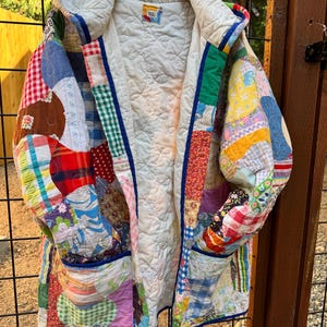 May include: A hooded jacket constructed from a patchwork of various colorful fabric scraps including gingham, floral, and plaid patterns. The garment features a white quilted interior lining, long sleeves, and two front pockets, all finished with blue trim.