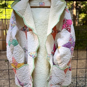 May include: Cream-colored quilted jacket with a hood, featuring patchwork designs in various colors like pink, orange, and green. The jacket has a light green trim and is hanging on a wooden hanger.