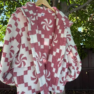 May include: A red and white patterned jacket hangs on a wooden hanger. The jacket features a checkerboard design with pinwheel-like patterns. The jacket is made of a soft, textured fabric and is hanging outside in front of a tree.