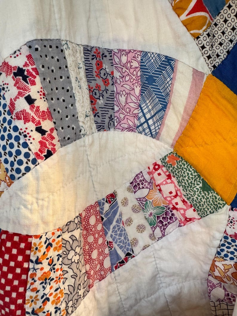 May include: Close-up of a vintage quilt with a white background and colorful, patterned fabric strips. The strips are arranged in a circular pattern, featuring floral, geometric, and abstract designs in red, blue, pink, and yellow.