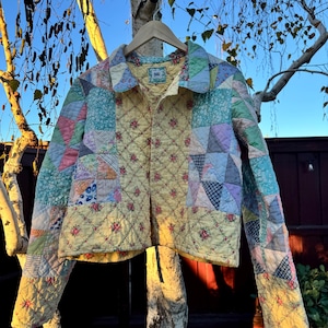 May include: A patchwork jacket with a variety of pastel-colored fabric squares and floral patterns. The jacket is hanging on a wooden hanger, showcasing its quilted design and button-up front. The jacket is a cropped style.