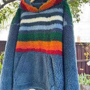 May include: A blue fleece hoodie with a striped design in shades of blue, white, green, orange, and red. The hood is trimmed with orange and red fleece. The hoodie has a kangaroo pocket and is hanging on a wooden hanger.