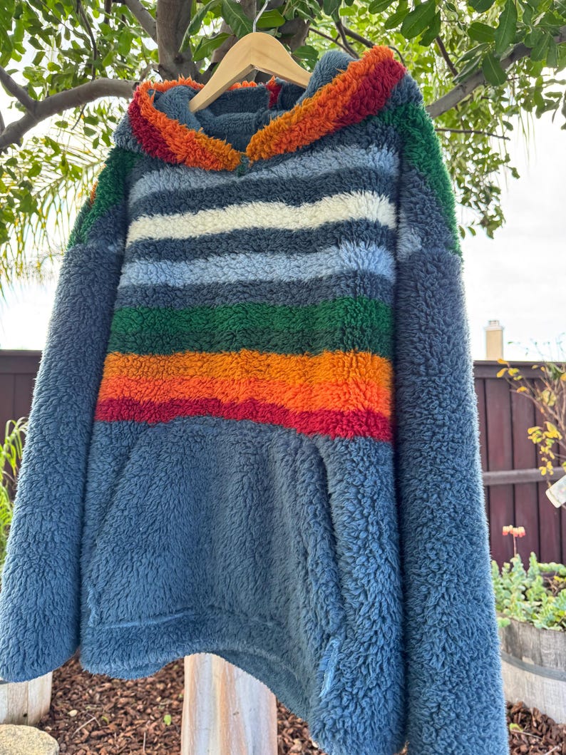 May include: A blue fleece hoodie with a striped design in shades of blue, white, green, orange, and red. The hood is trimmed with orange and red fleece. The hoodie has a kangaroo pocket and is hanging on a wooden hanger.