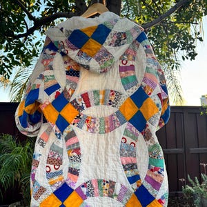 May include: A handmade, quilted jacket with a hood, featuring a patchwork design. The jacket showcases a mix of colorful fabrics in a variety of patterns, including blue, yellow, and patterned fabrics. The jacket is hanging on a wooden hanger.