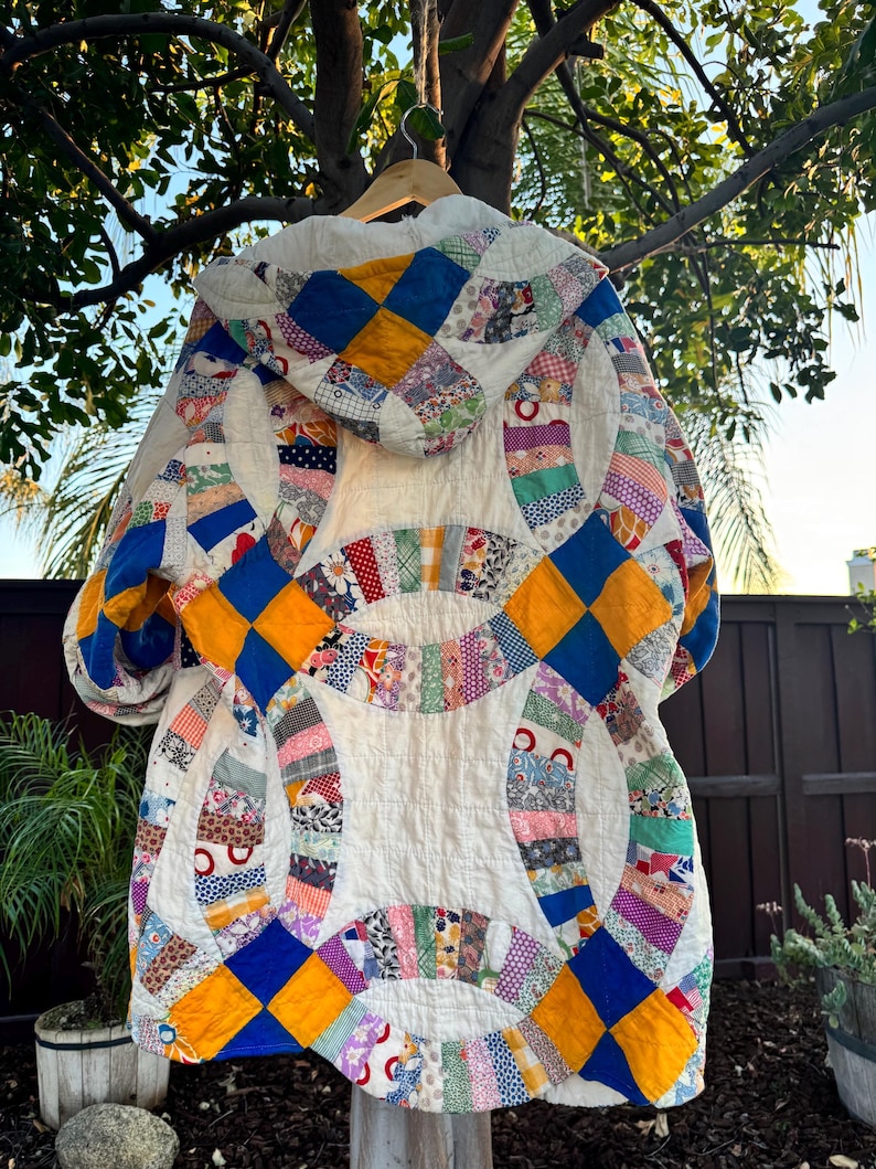 May include: A handmade, quilted jacket with a hood, featuring a patchwork design. The jacket showcases a mix of colorful fabrics in a variety of patterns, including blue, yellow, and patterned fabrics. The jacket is hanging on a wooden hanger.