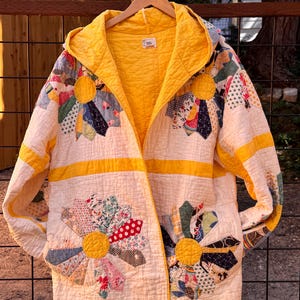 May include: A hooded jacket crafted from a quilted fabric featuring a cream base with bright yellow trim. The garment displays large, multi-colored floral patterns made of various fabric scraps, including polka dots and stripes, arranged in a fan-like design.