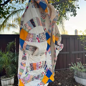 May include: A handmade quilt coat with a patchwork design. The coat features a variety of colorful fabric scraps in a mix of patterns, including floral, geometric, and solid colors. The coat is hanging on a wooden hanger outdoors.